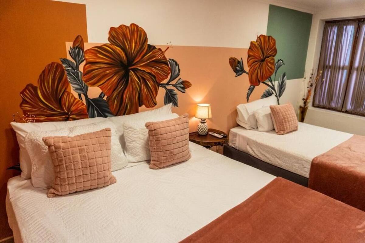Habitación de hotel con dos camas, una mesita de noche con lámpara y un gran mural floral en la pared.
