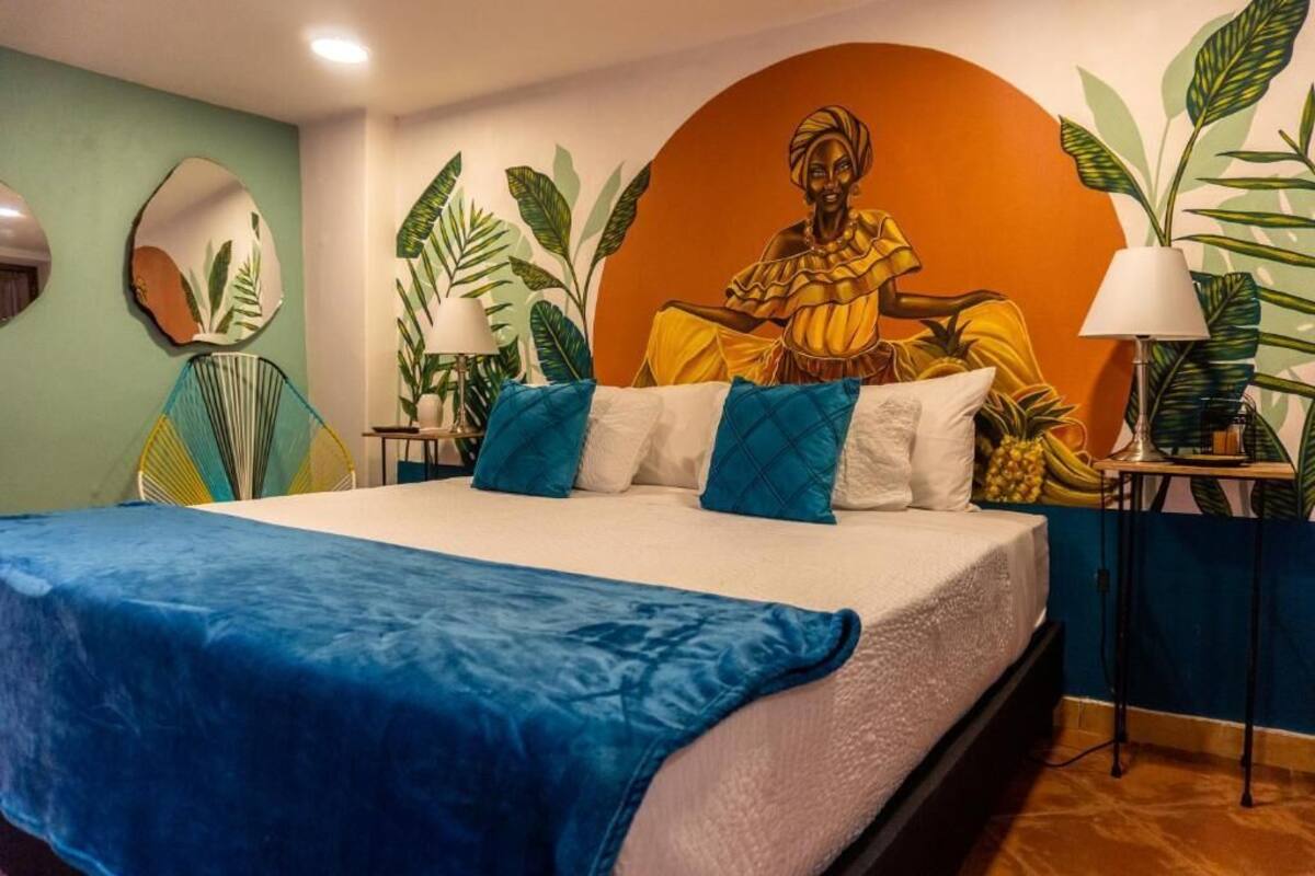 Una cama con una manta azul, almohadas blancas y un mural tropical en la pared.