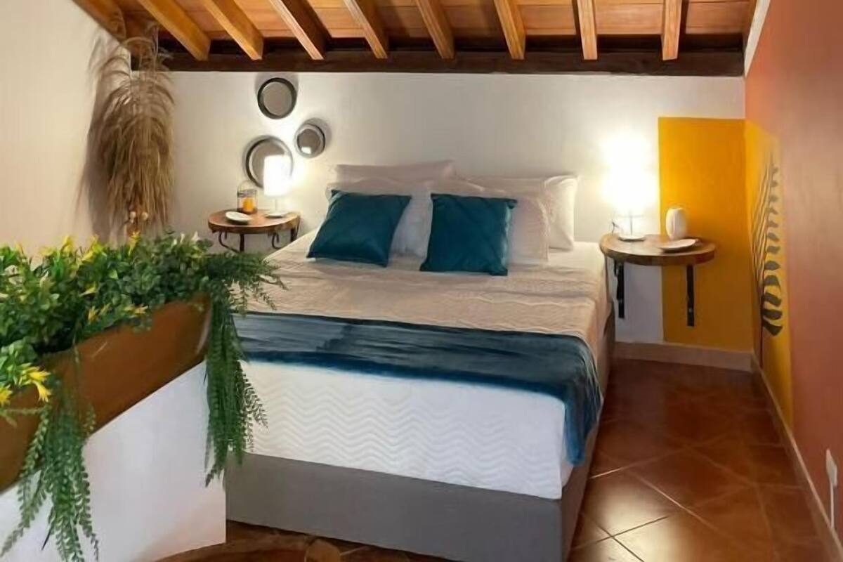Un dormitorio con techo de madera, una cama con ropa de cama azul y blanca, una mesita de noche con lámpara y un espejo fijado a la pared.