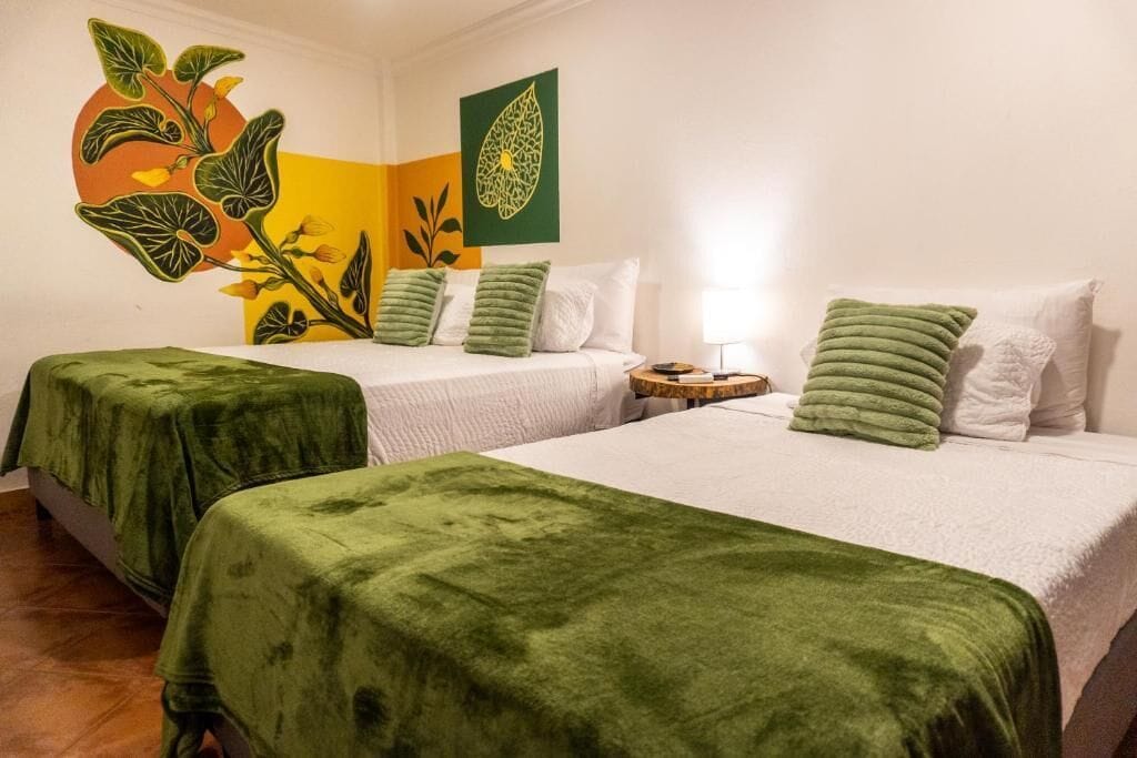 A hotel room with two beds, green bedspreads, white bedding, a nightstand, and a wall with a plant mural.