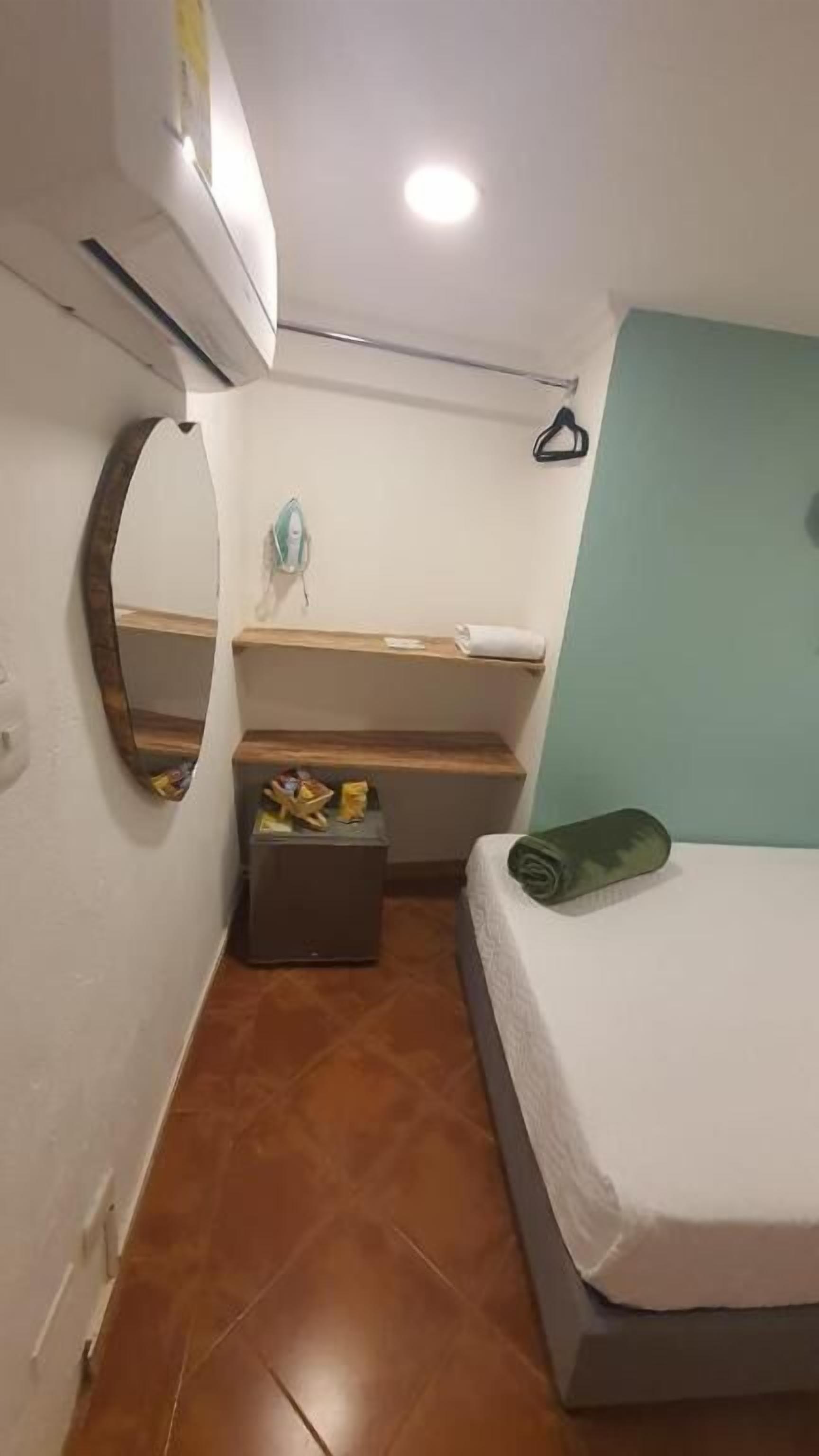 A small, compact room with a bed, a mirror, a shelf with toiletries, and a hanging coat rack.