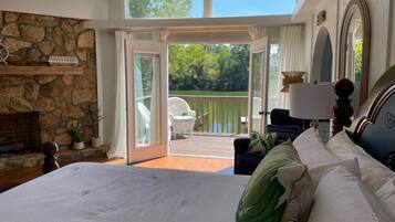 A bedroom with a large bed, a stone fireplace, a balcony with a chair, and a view of the outdoors.