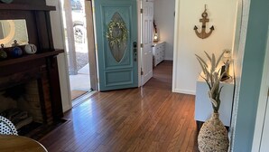 Un couloir au plancher de bois, avec une porte de couleur turquoise, un mur blanc orné d’une ancre et un vase en osier contenant des plantes.