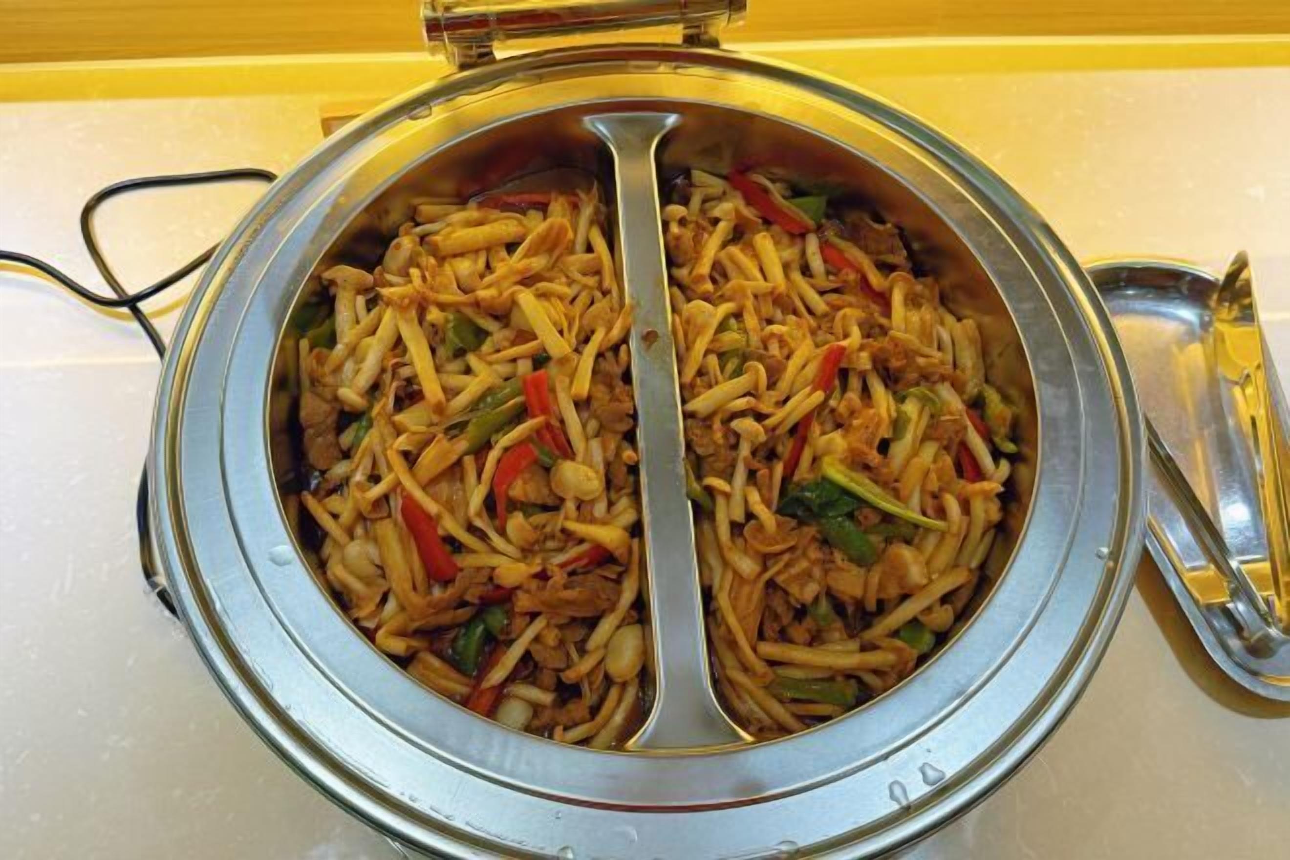 Recipiente metálico redondo com uma colher, cheio de um prato de macarrão com legumes.