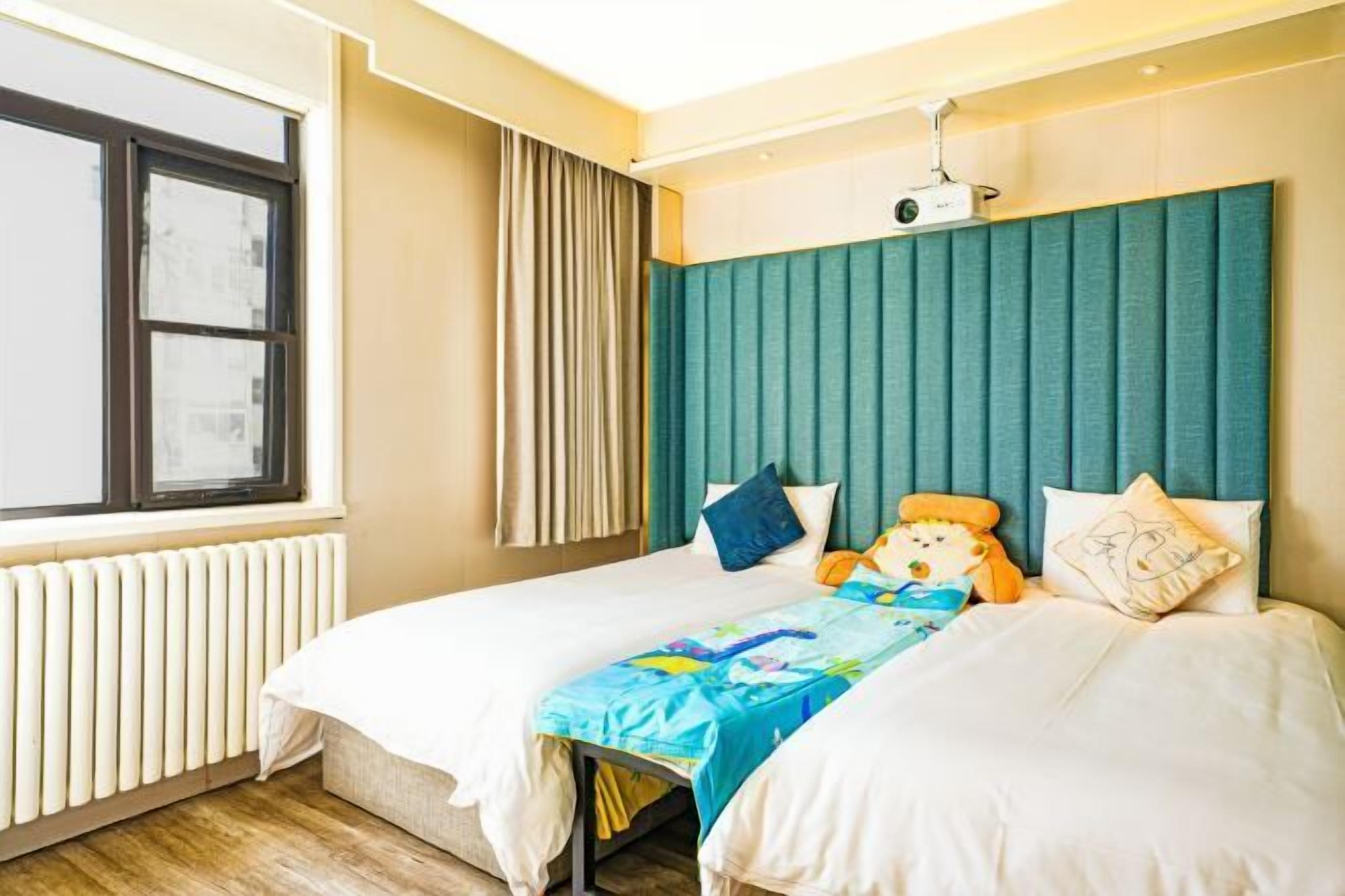 A hotel room with two beds, a window, a radiator, and a projector on the ceiling.