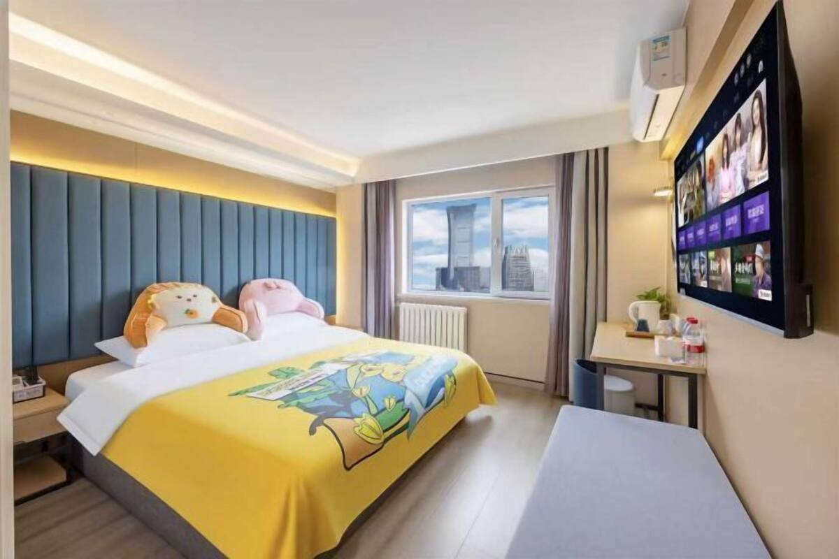 A hotel room with a bed, a desk, a chair, a window with a city view, and a wall-mounted TV.