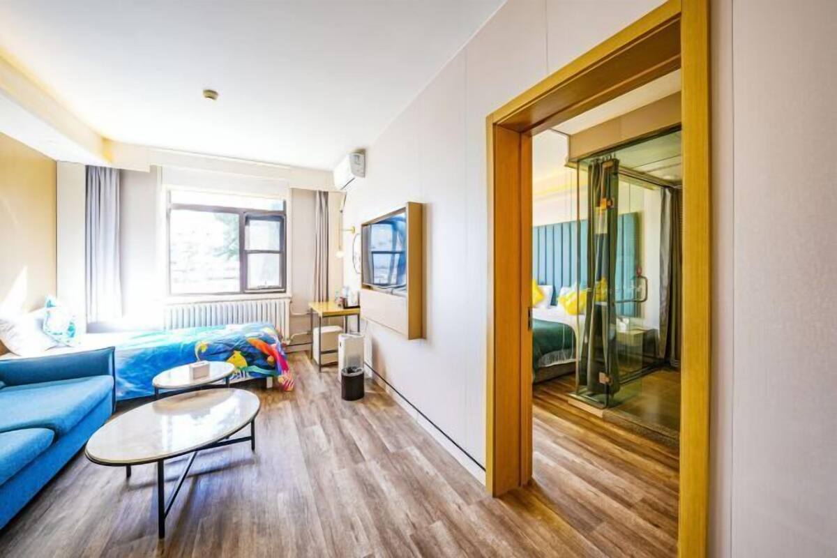 A modern hotel room with a wooden floor, a blue sofa, a small table, a glass partition, and a view of the outdoors.