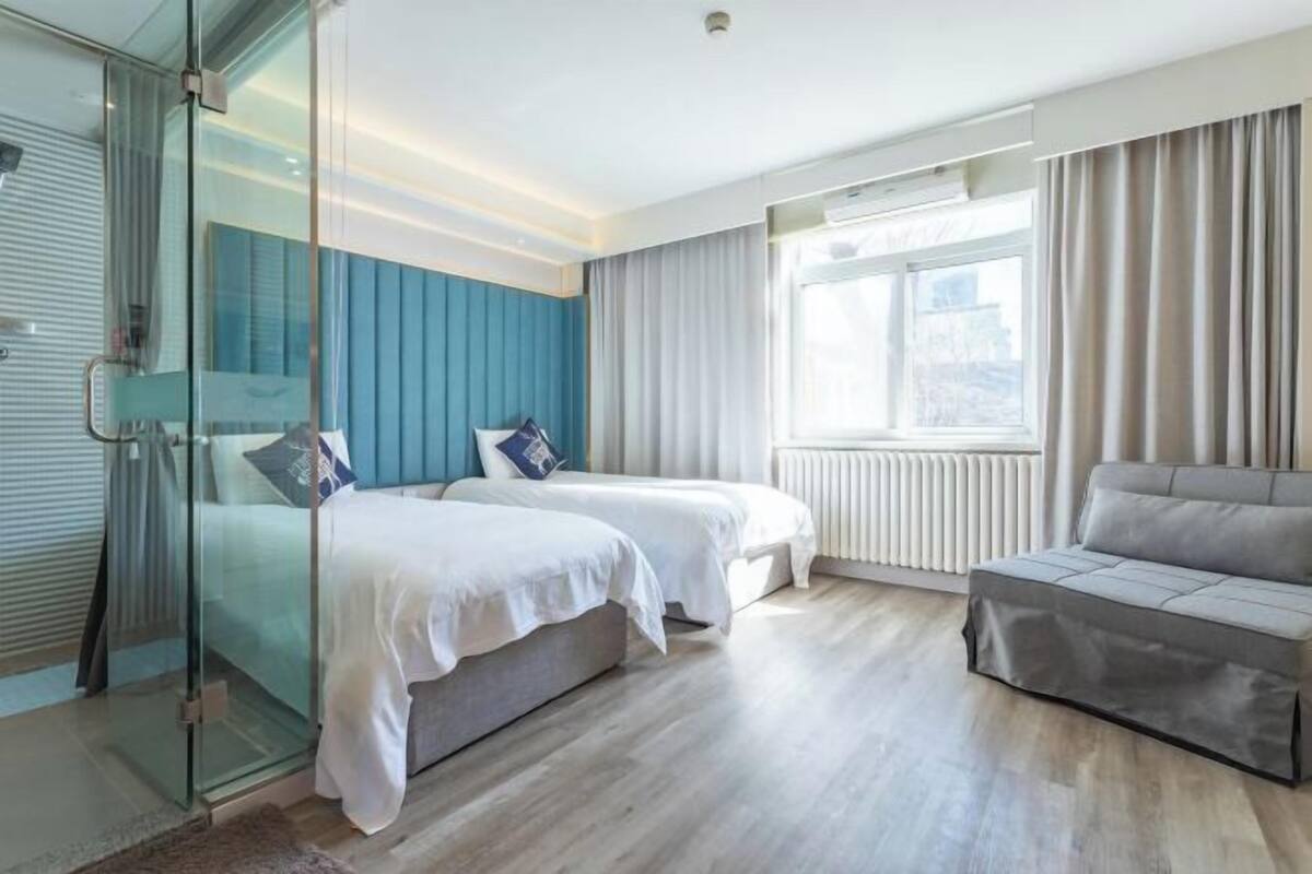 A modern hotel room with two beds, a sofa, a glass shower enclosure, and a large window with curtains.