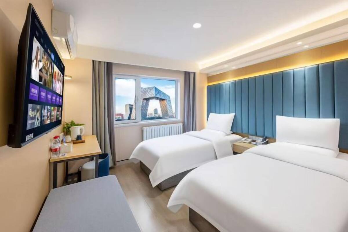 A hotel room with two beds, a desk with a computer, a TV mounted on the wall, and a window with a cityscape view.