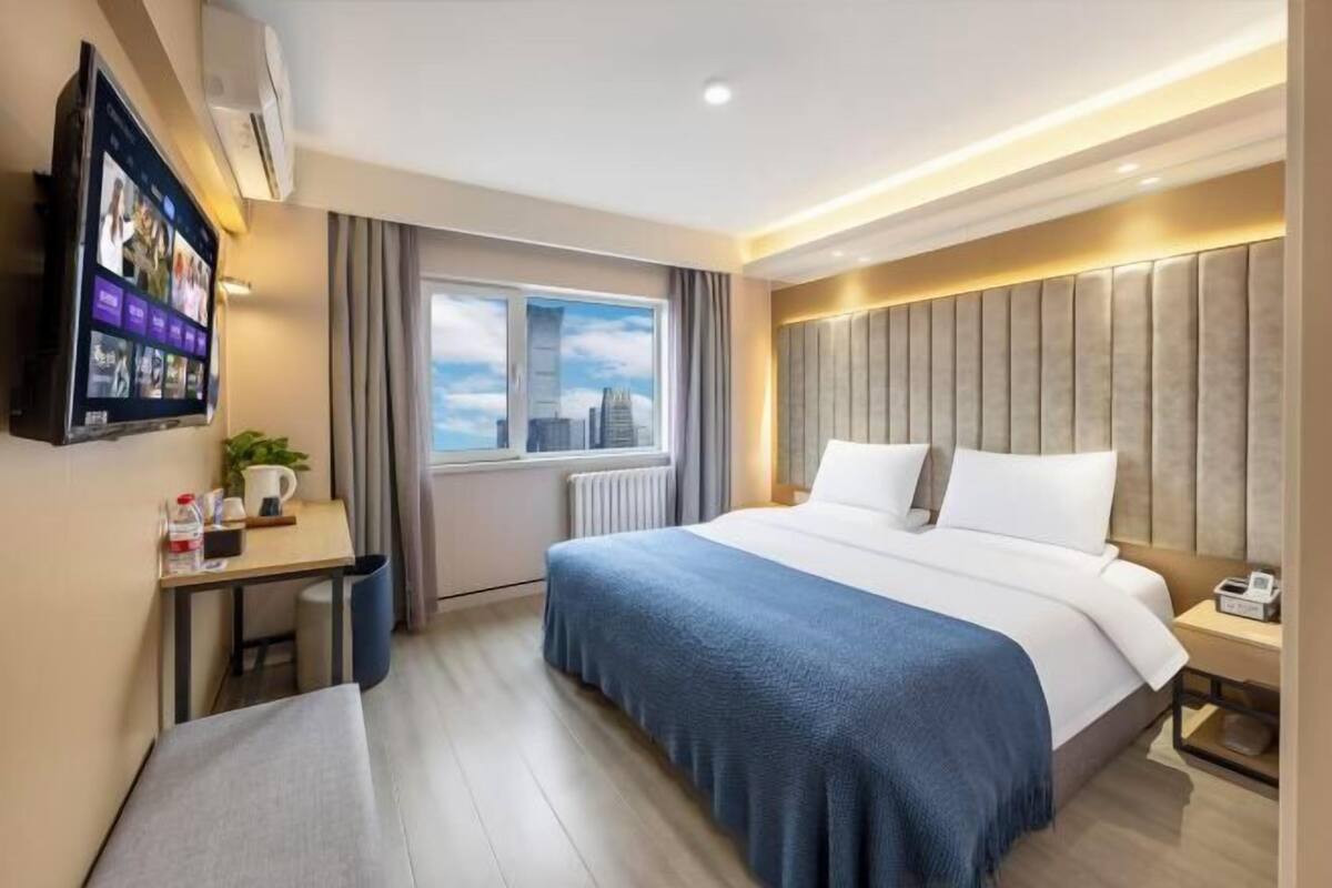 A hotel room with a bed, a desk, a television, and a window with a city view.