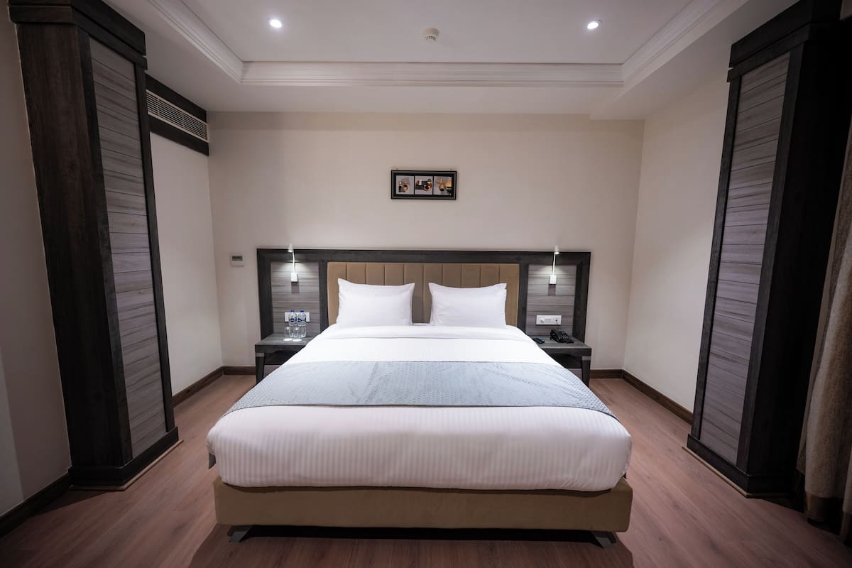 A spacious hotel room with a large bed, two bedside tables, a headboard with integrated lighting, and a framed picture on the wall.
