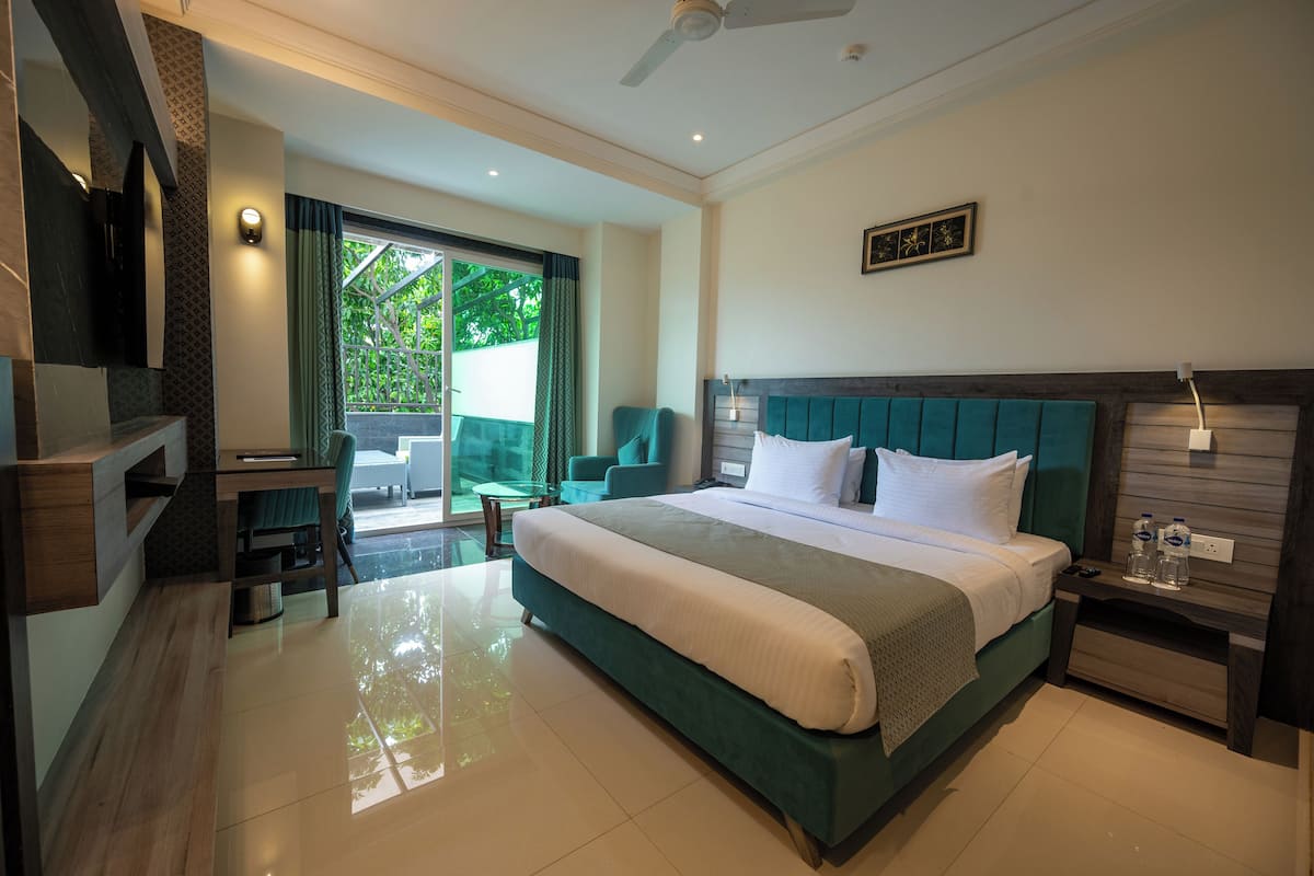 A modern hotel room with a large bed, a TV, a desk, and a balcony with a view of greenery.