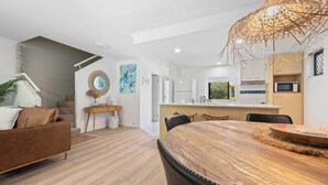 A modern kitchen with white cabinets, a microwave, and a dining area with a wooden table and chairs. - Unit 94 - Modern and Stylish 2 Bedroom Townhouse in Resort (Noosaville)