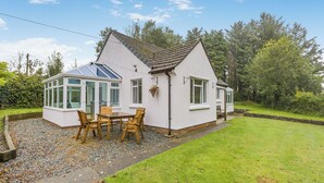 A white house with a conservatory, a patio with wooden furniture, and a well-maintained garden. - Pine Cottage - UK46874 (Hartland)