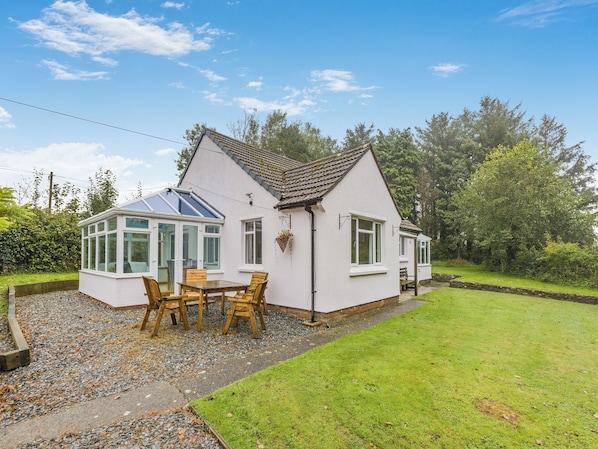 A white house with a conservatory, a patio with wooden furniture, and a well-maintained garden.