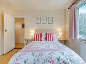 A bedroom with a floral bedspread, two bedside lamps, a window with curtains, and a door leading to another room.