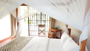 A bedroom with a bed, a small table, and a chair. There is a window with a view of greenery outside. - Ilboru Safari Lodge - The Platinum Route Company Limited (Arusha)