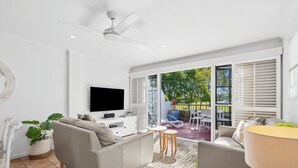 A modern living room with a flat-screen TV, grey sofas, a ceiling fan, and a view of the outdoors through large windows.