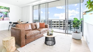 Un salon moderne avec un canapé en cuir marron, une table basse ronde et une grande porte-fenêtre coulissante donnant sur un balcon d’où l’on peut admirer le ciel et les palmiers.