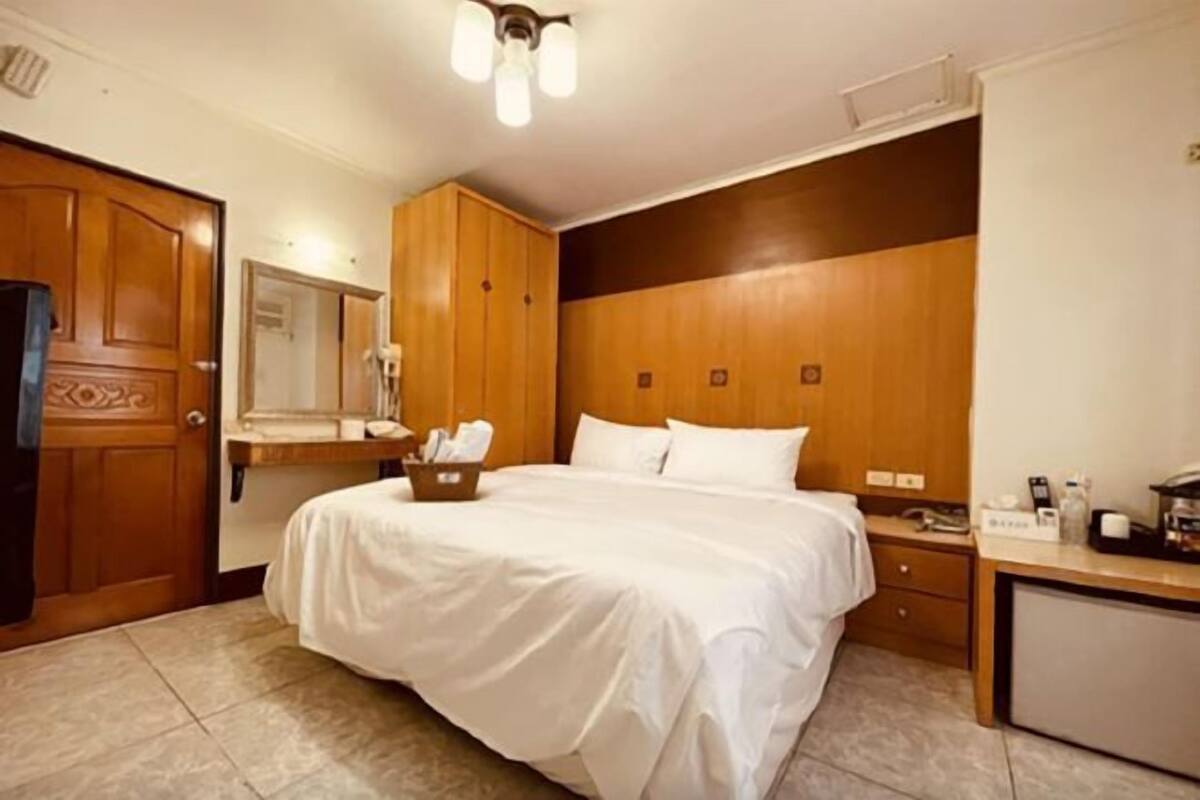 A hotel room with a large bed, wooden wardrobe, a desk with a chair, and a small kitchenette.