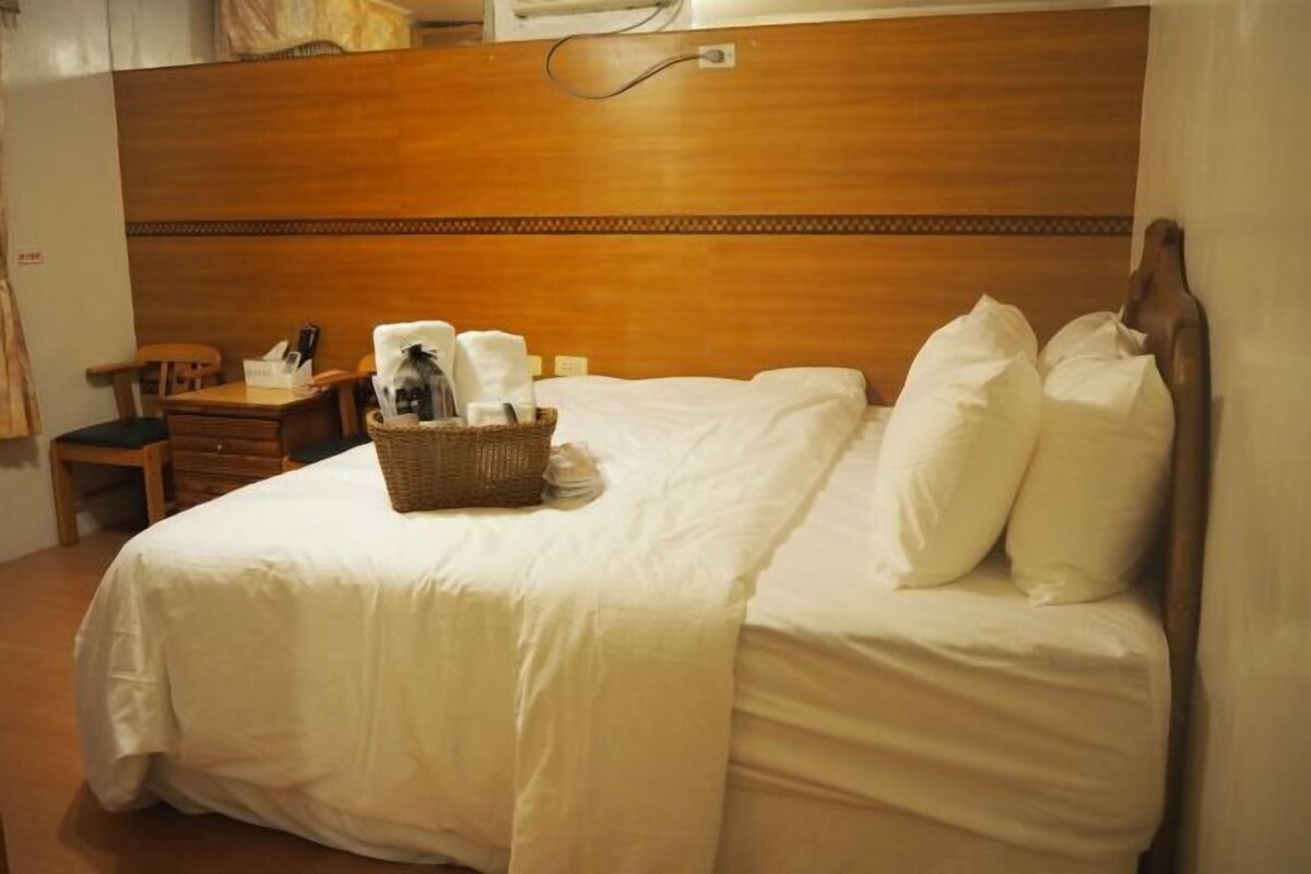 A hotel room with a wooden headboard, a neatly made bed with white linens, a small wooden nightstand with a lamp and a basket of toiletries.