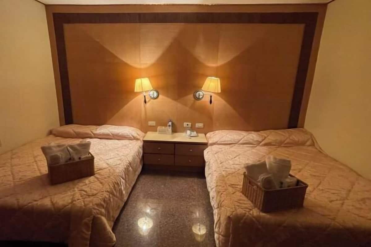 A hotel room with two beds, a small desk, and two wall-mounted lamps.