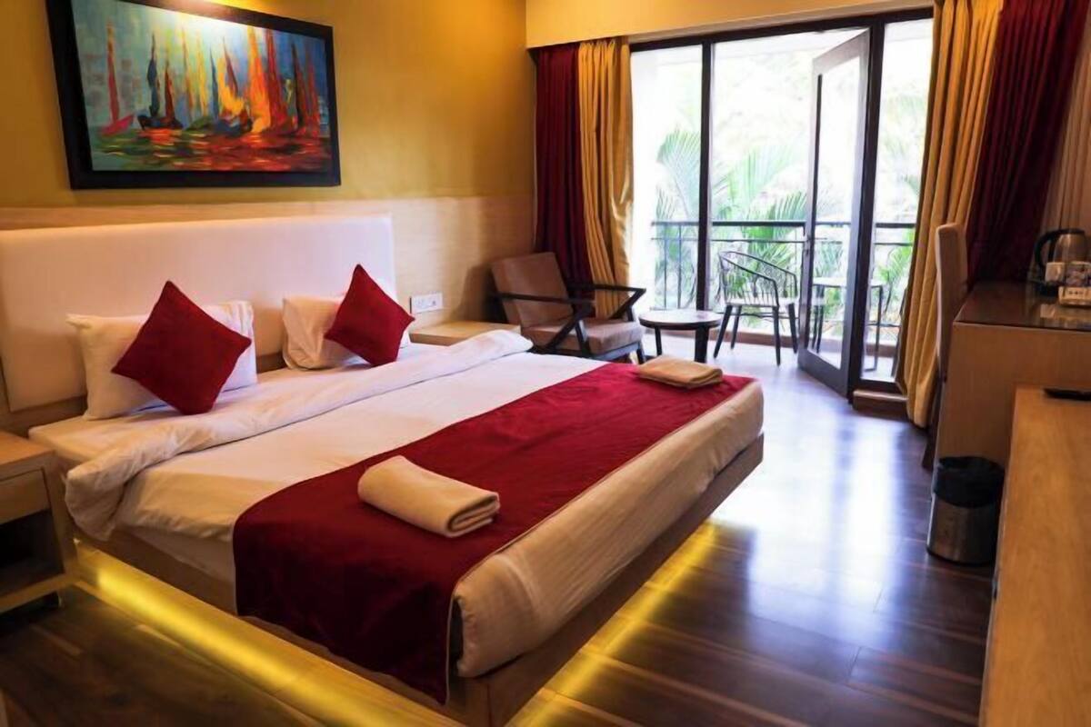 A hotel room with a large bed, a painting on the wall, a balcony with chairs, and a desk.