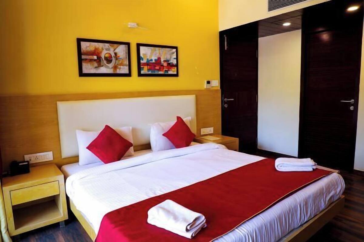 A hotel room with a large bed, two red pillows, a red throw blanket, and a white towel on the bed. There are two paintings on the wall above the bed.
