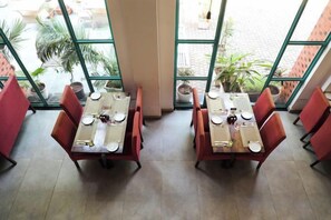 Un restaurante con mesas dispuestas para comer, amplios ventanales con vista a la calle y plantas en macetas.
