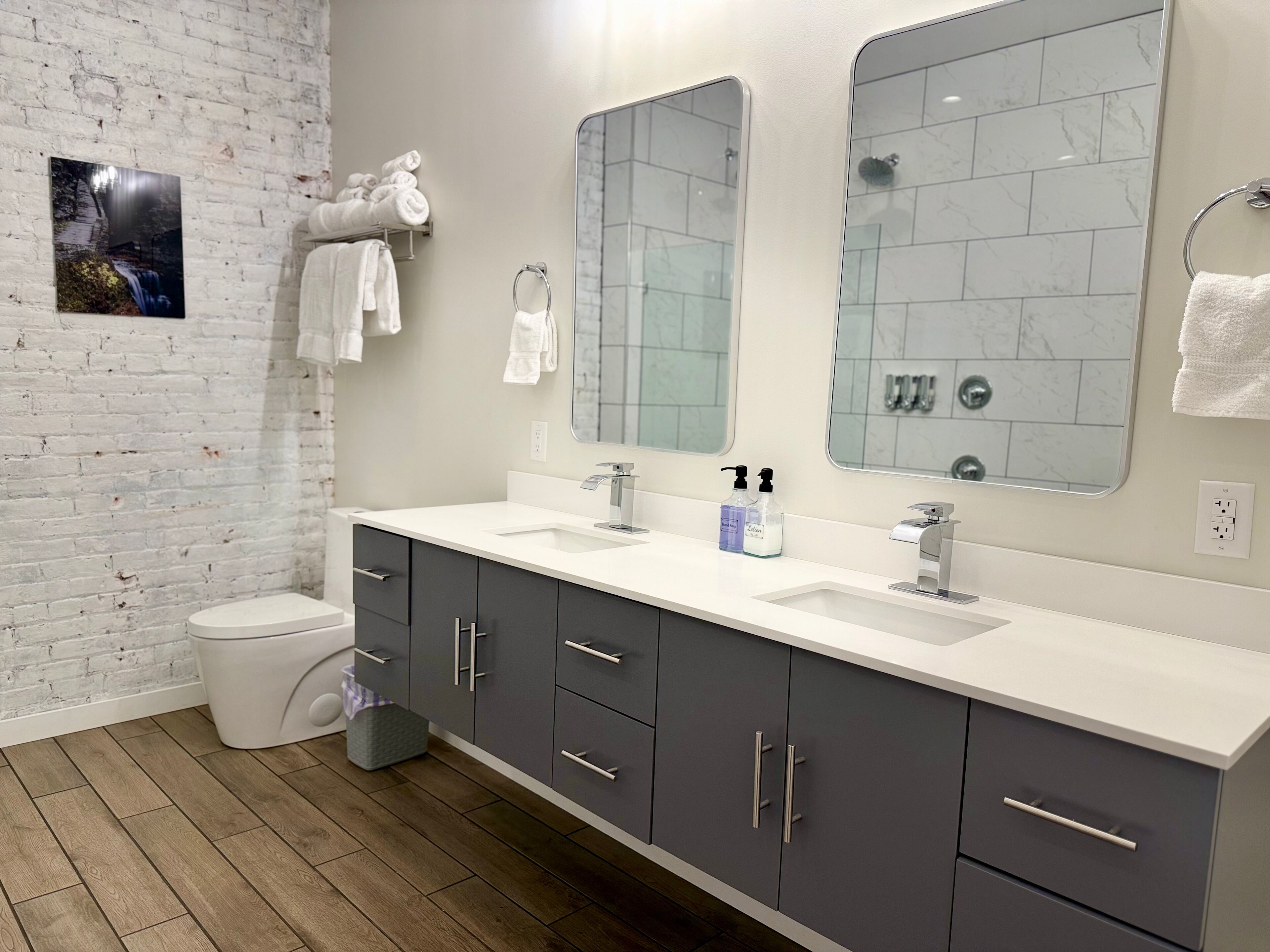 Un baño moderno con pared de ladrillo blanco, dos lavamanos, armarios grises oscuros y un espejo grande.