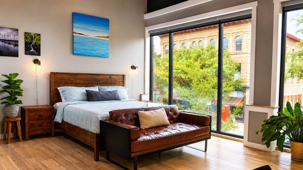 Un dormitorio con una cama grande, un sofá de cuero, piso de madera y vista a un edificio a través de amplios ventanales.