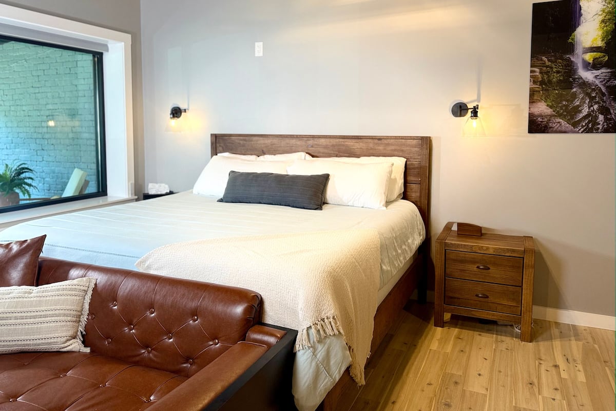 Un dormitorio con una cama grande, una mesita de noche de madera, un sofá de cuero marrón y una ventana con vistas a un muro de ladrillos y vegetación.