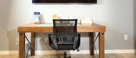 A room with a large flat-screen TV mounted on the wall, a wooden desk with a black office chair, and two framed pictures on the wall.