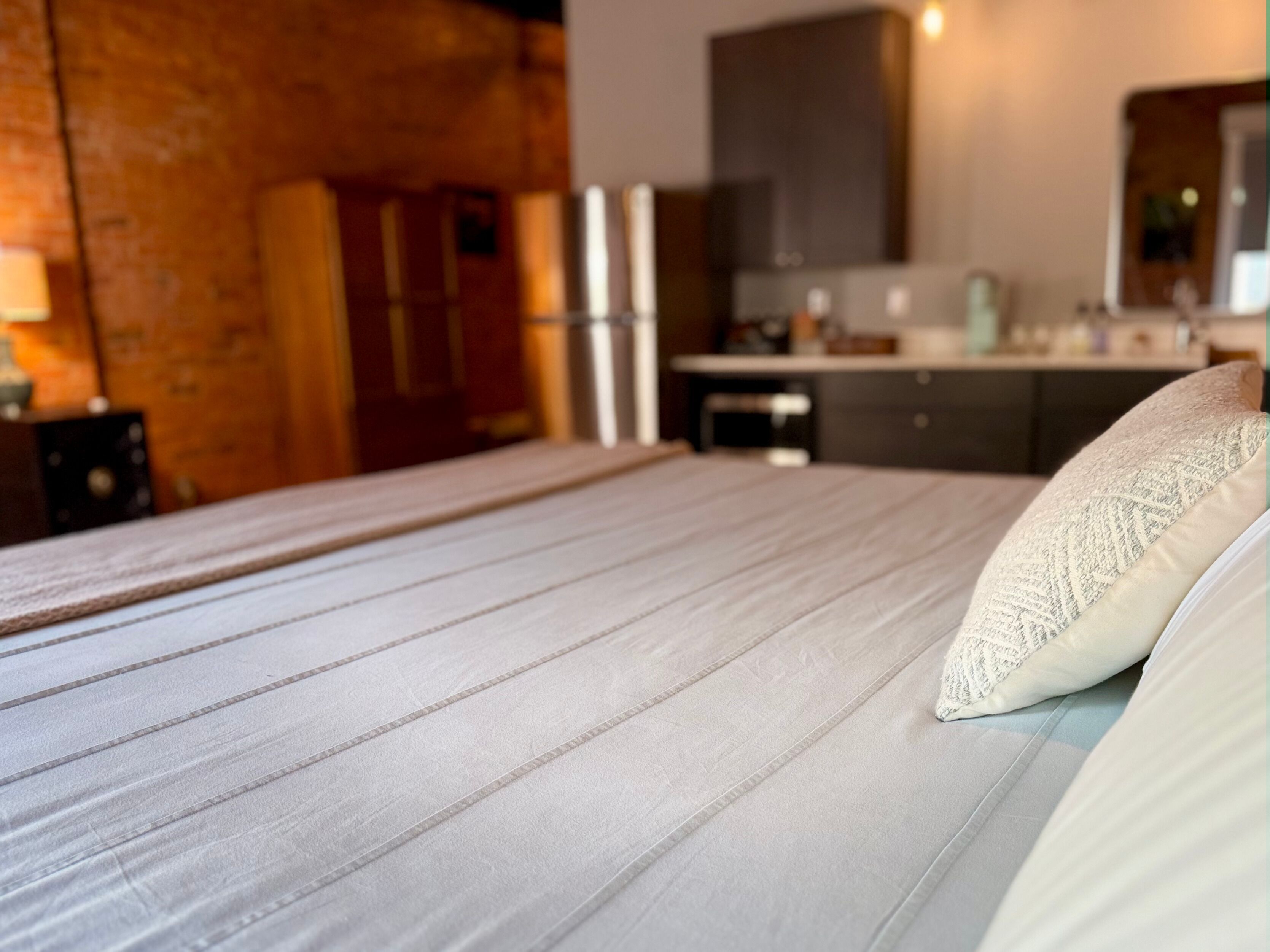 Un llit ben arropat amb una almoinada en una habitació amb paret de fusta i una cuina al fons.
