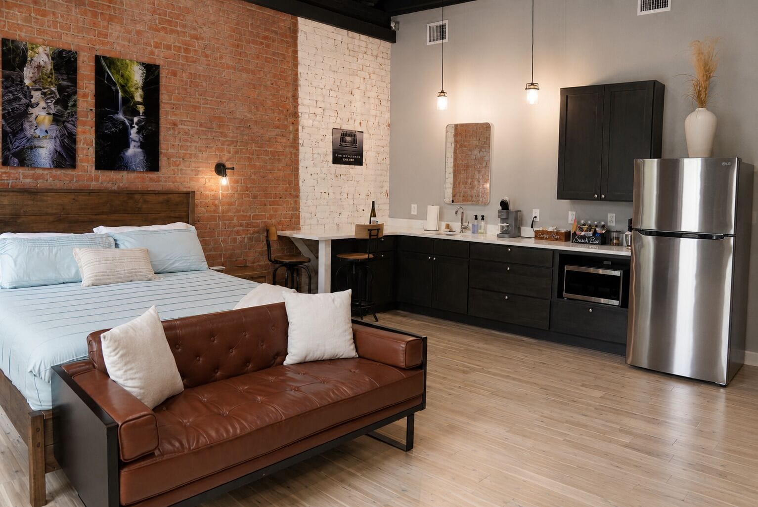 Un dormitorio moderno con una cama grande, un sofá de cuero marrón y una cocina con armarios oscuros y un refrigerador de acero inoxidable.