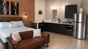 Un dormitorio moderno con una cama grande, un sofá de cuero marrón y una cocina con armarios oscuros y un refrigerador de acero inoxidable.