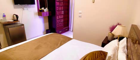 Habitación de hotel con una cama, un televisor montado en la pared, un equipo de aire acondicionado, un minibar y una mesita con una tetera y una botella.