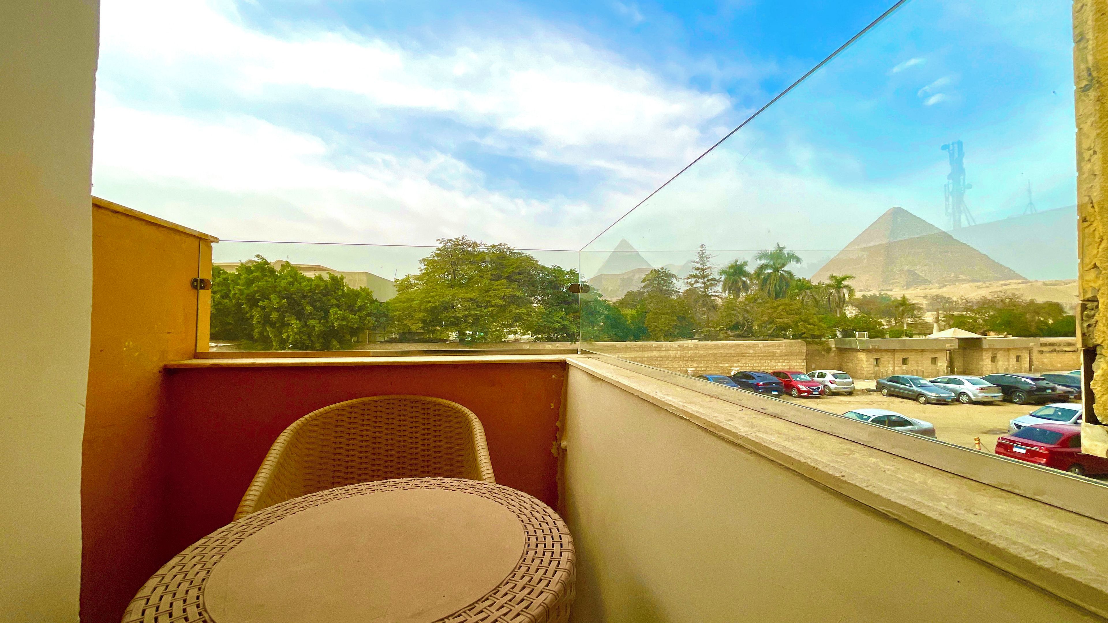 Een balkon met een rieten stoel, uitzicht op een parkeerplaats en een piramide in de verte.