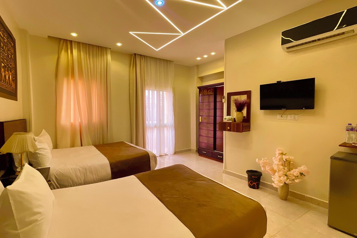 A hotel room with two beds, a flat-screen TV, a minibar, and a large window with curtains.