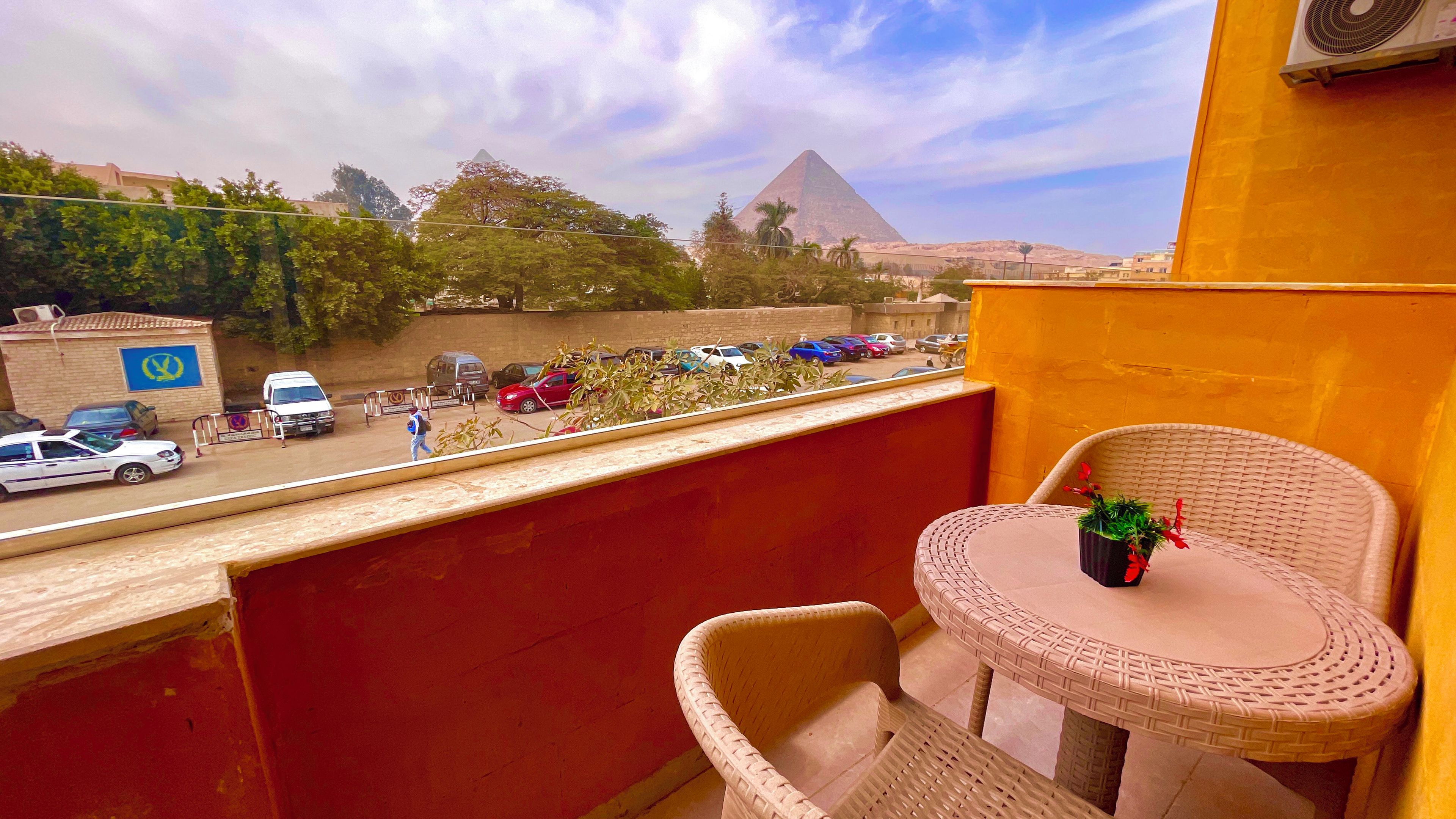 A balcony with wicker chairs and a small table, overlooking a parking area and a pyramid in the distance.