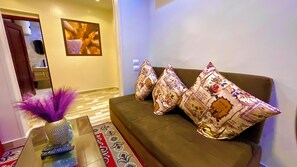 Una sala de estar moderna con un sofá marrón, cojines estampados, un florero decorativo con plumas moradas y un control remoto sobre una mesa de centro de vidrio.
