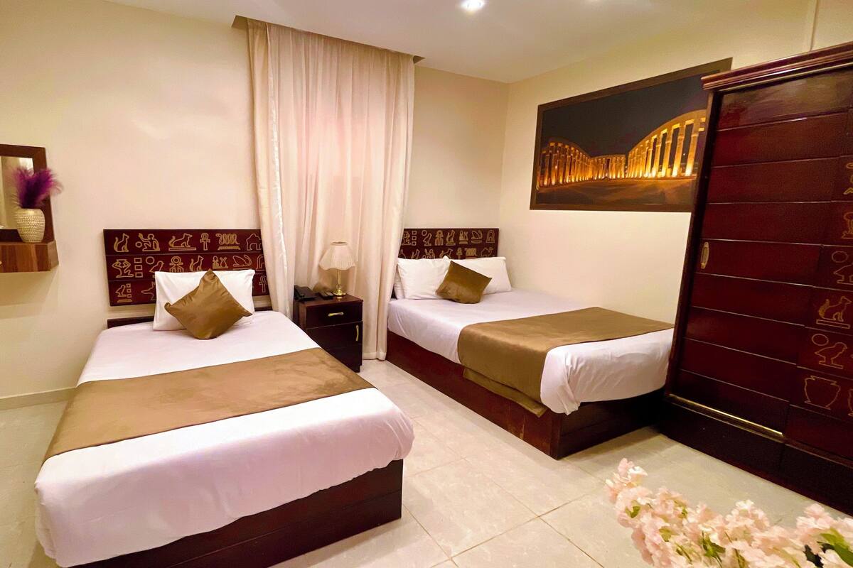 A hotel room with two beds, a nightstand, a mirror, a wall-mounted artwork, and a wardrobe.