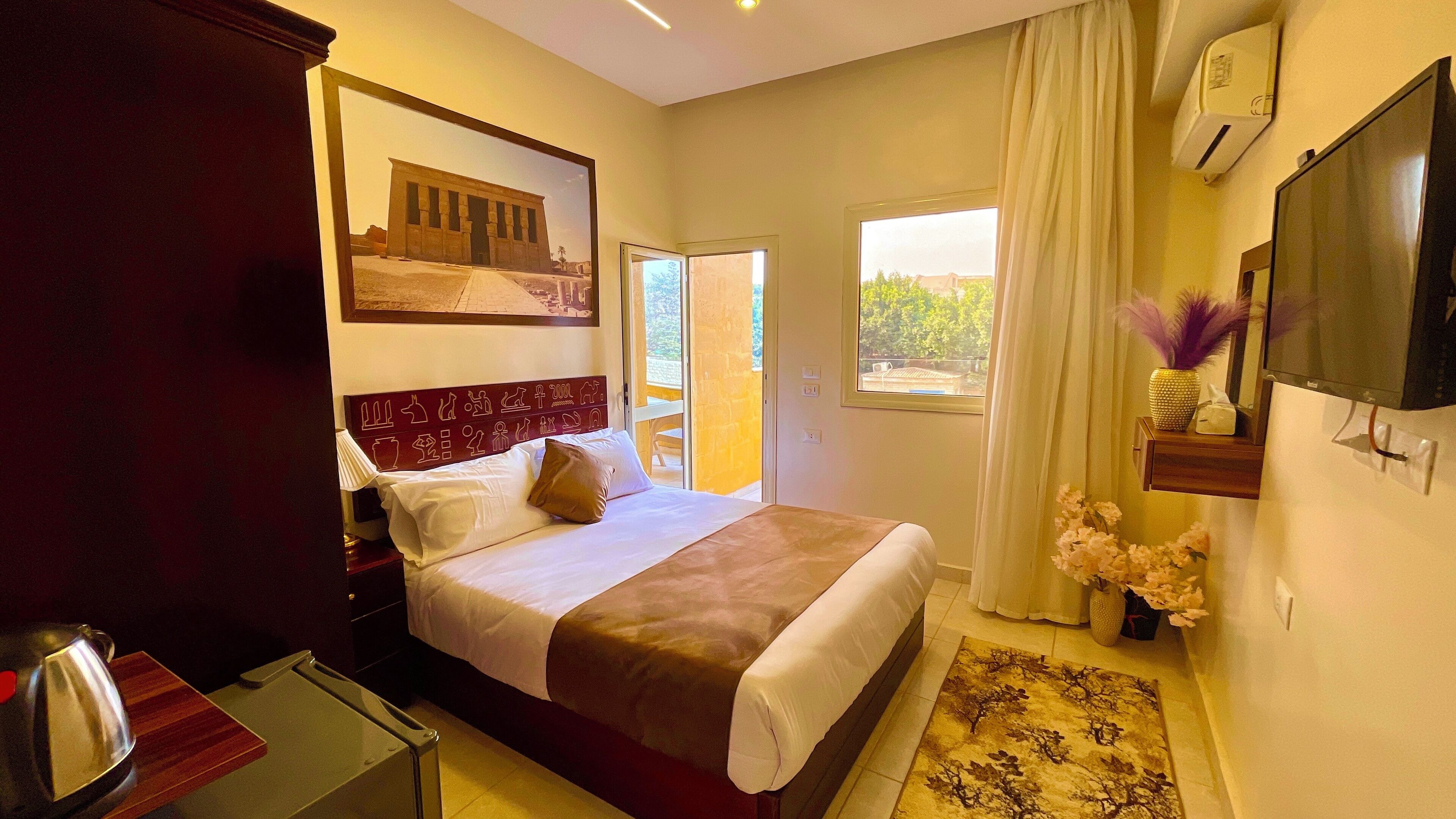 A hotel room with a bed, a flat-screen TV, a window with curtains, a small table with a lamp, and a decorative wall piece.