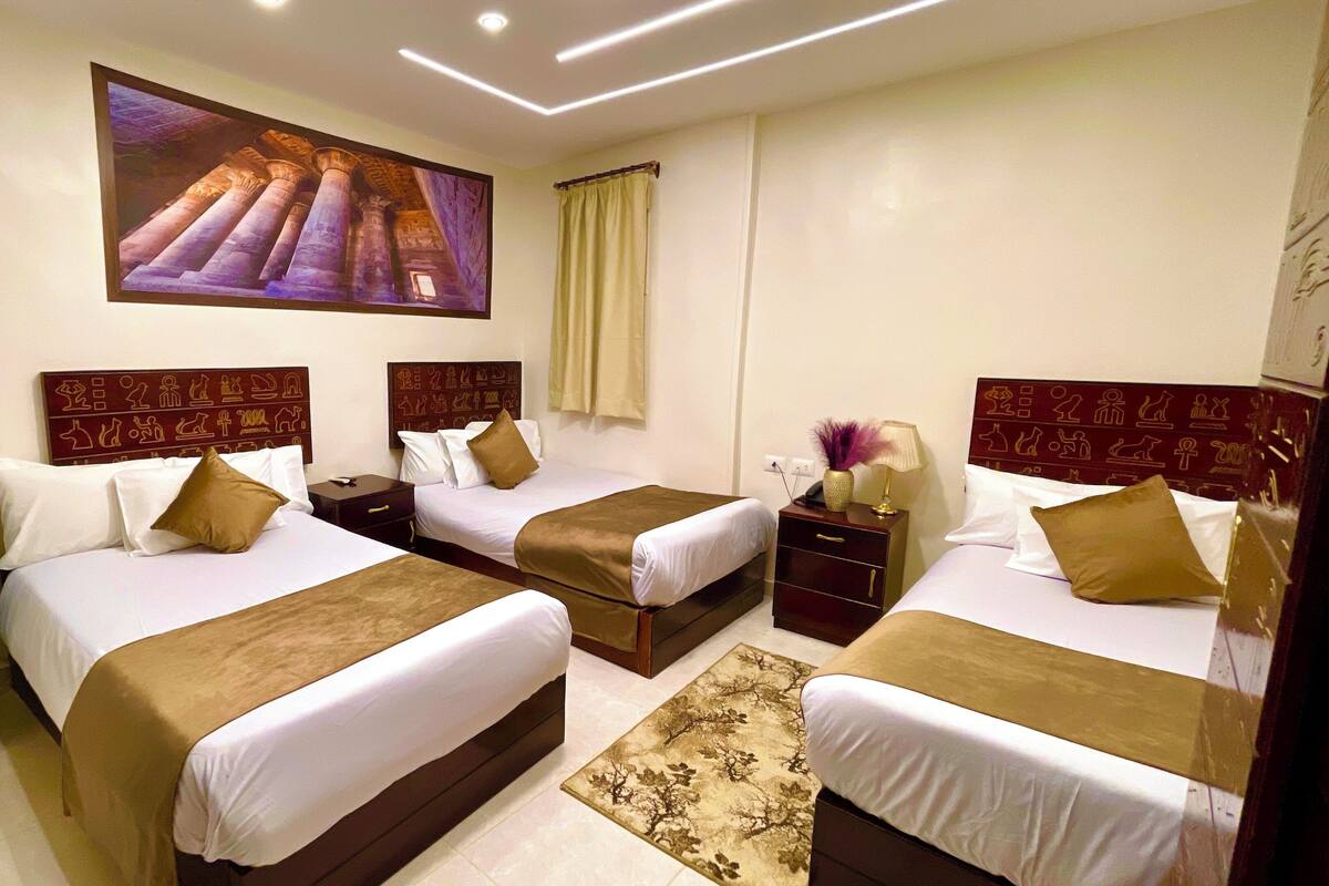 A hotel room with three beds, a large wall painting, a minibar, and a decorative rug.