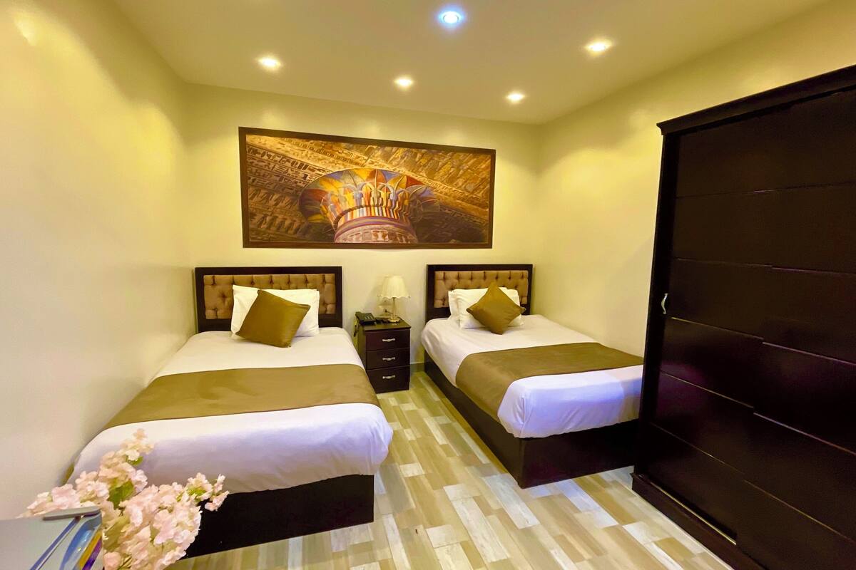 A hotel room with two beds, a large painting above the beds, a desk with a chair, and a wardrobe.