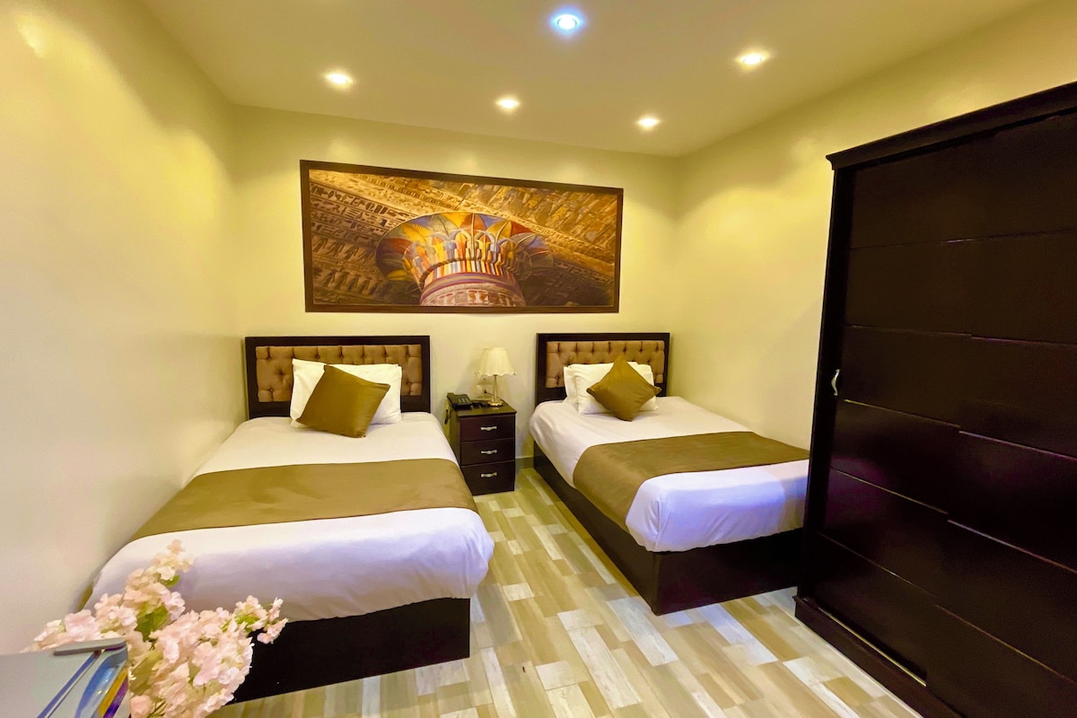 A hotel room with two beds, a large painting above the beds, a desk with a chair, and a wardrobe.