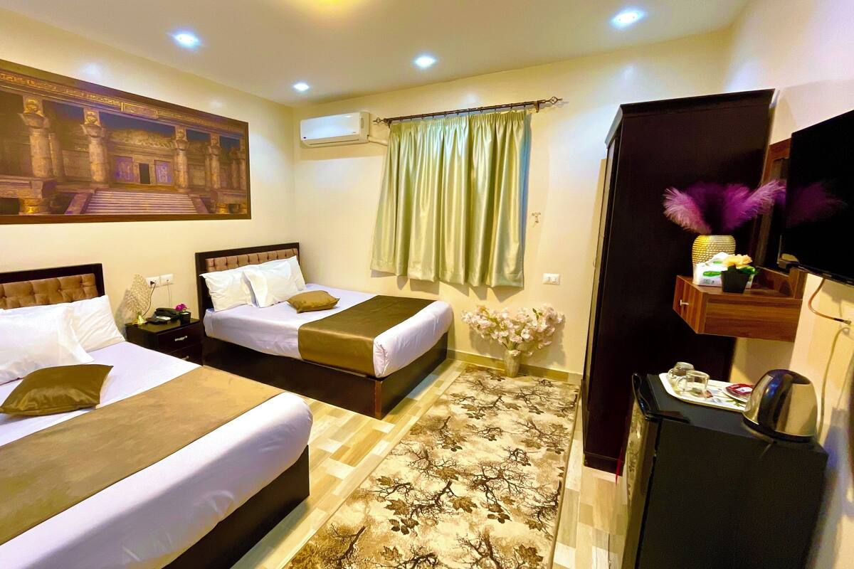 A hotel room with two beds, a large painting on the wall, an air conditioning unit, a decorative vase, and a small table with a remote control.