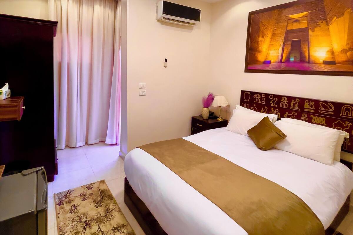 A hotel room with a large bed, an air conditioning unit, a painting on the wall, and a desk with a lamp.
