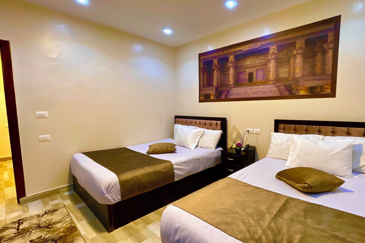 A hotel room with two beds, a nightstand, a lamp, and a painting on the wall.