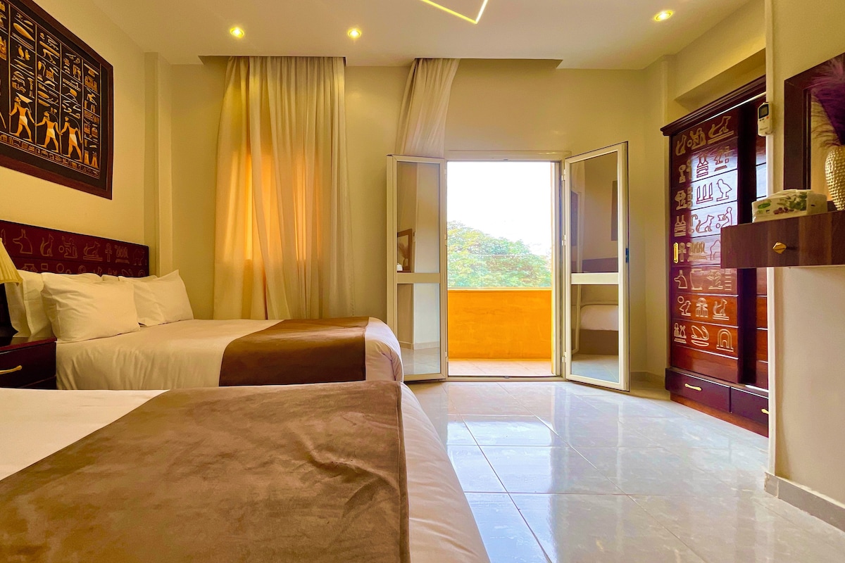 A hotel room with two beds, a large window with curtains, a wooden headboard, and a decorative wall piece.